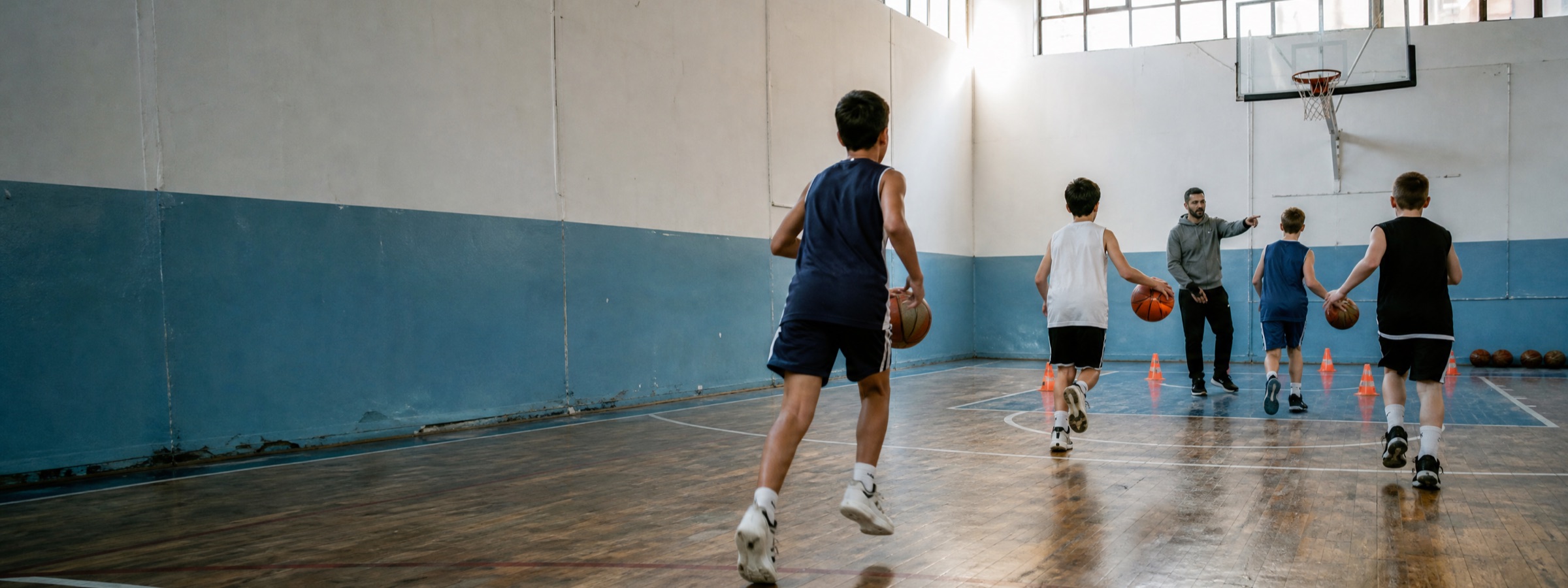 Mahalle spor salonunda basketbol antrenmanı; antrenör bir grup çocuğa hareket gösteriyor